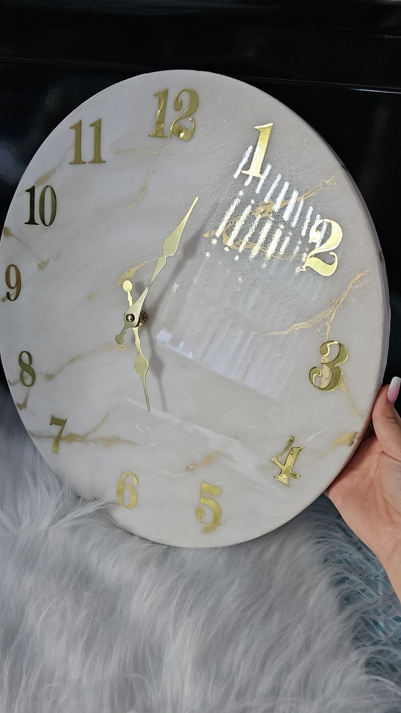 May include: A white and gold marble clock with gold Roman numeral hour markers and gold hands. The clock face has a white background with gold marble accents.