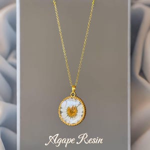 May include: A gold necklace with a circular pendant containing a preserved daisy flower in clear resin. The pendant has a gold-coloured border. The text "Agape Resin" is printed on the grey box.