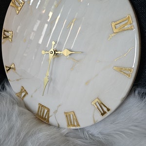 May include: White round clock with gold Roman numeral markings and gold hands. The clock face has a marble-like design with gold streaks.