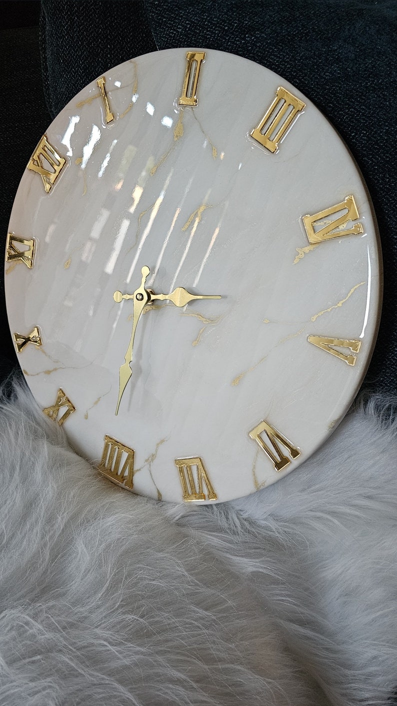 May include: White round clock with gold Roman numeral markings and gold hands. The clock face has a marble-like design with gold streaks.