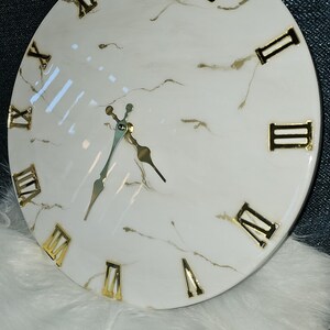 May include: A white round clock with gold Roman numeral markings and gold hands. The clock face has a marble-like design.