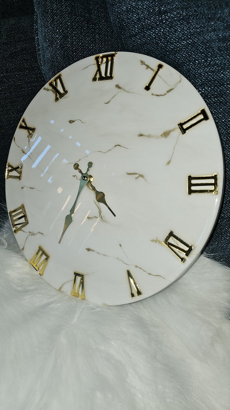 May include: A white round clock with gold Roman numeral markings and gold hands. The clock face has a marble-like design.