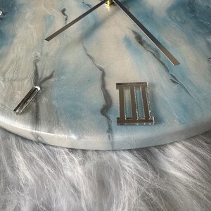 Puede incluir: Un reloj de resina blanco y azul con manecillas plateadas y n&uacute;meros romanos. El reloj est&aacute; colocado sobre un fondo blanco y esponjoso.