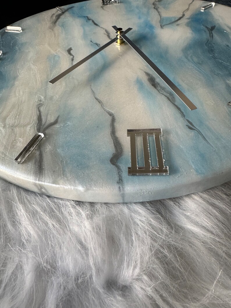 Puede incluir: Un reloj de resina blanco y azul con manecillas plateadas y n&uacute;meros romanos. El reloj est&aacute; colocado sobre un fondo blanco y esponjoso.