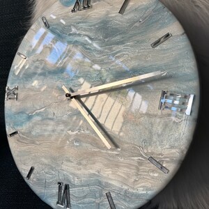 Puede incluir: Un reloj redondo, blanco y azul con n&uacute;meros romanos plateados y manecillas plateadas. La esfera del reloj tiene un dise&ntilde;o marmoleado y abstracto.