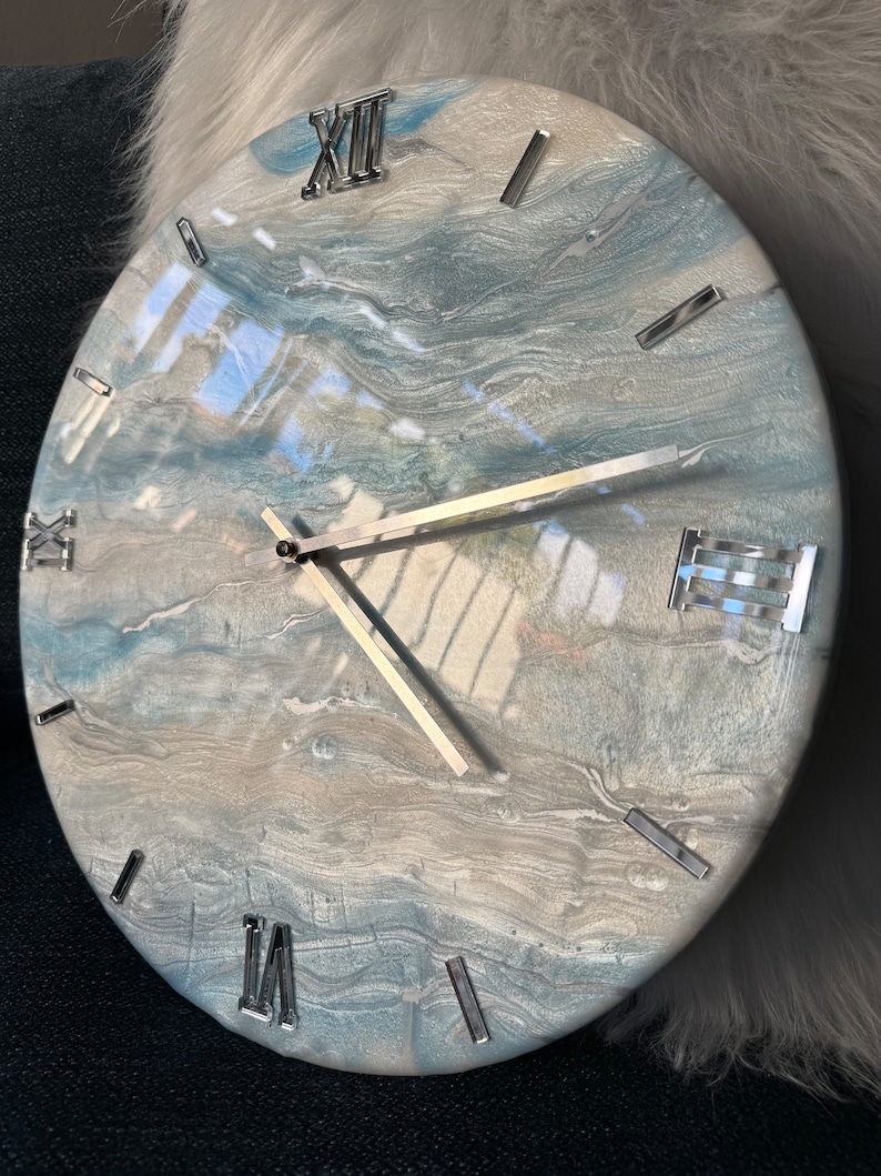 Puede incluir: Un reloj redondo, blanco y azul con n&uacute;meros romanos plateados y manecillas plateadas. La esfera del reloj tiene un dise&ntilde;o marmoleado y abstracto.