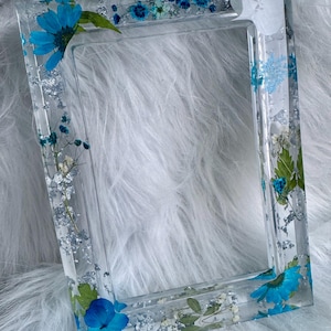 May include: A rectangular clear resin picture frame featuring embedded blue flowers, green leaves, and silver leaf flakes. The frame has a smooth, glossy finish with a hollow center, showcasing a variety of dried floral elements throughout the structure.