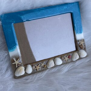 May include: A rectangular picture frame featuring a glossy blue and white resin top section resembling ocean waves. The bottom section is textured with a sandy appearance, decorated with various white seashells and small tan starfish.