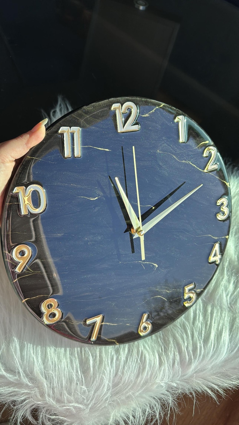 May include: A black and gold clock with a marble effect. The clock face has white Roman numerals and gold hands.