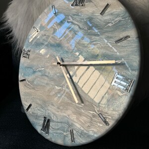 Puede incluir: Un reloj redondo blanco con acentos marmoleados azules y grises. La esfera del reloj tiene marcas de n&uacute;meros romanos y manecillas plateadas.