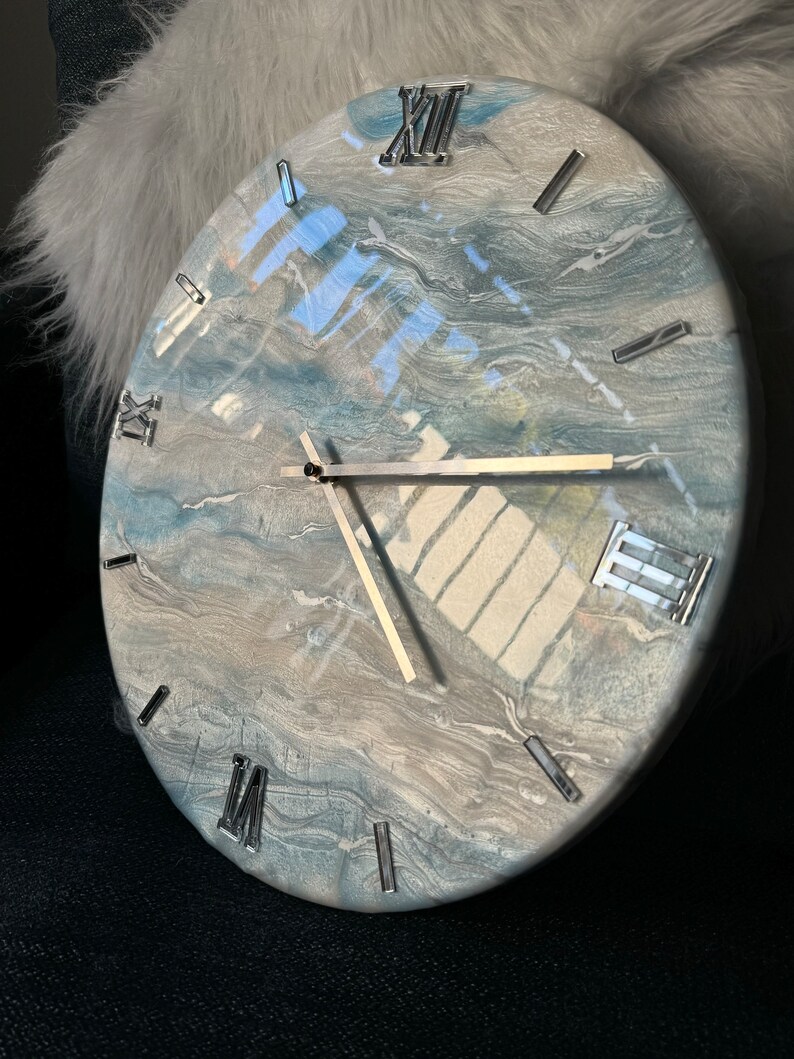 Puede incluir: Un reloj redondo blanco con acentos marmoleados azules y grises. La esfera del reloj tiene marcas de n&uacute;meros romanos y manecillas plateadas.