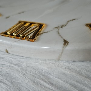 May include: A white marble clock face with gold Roman numeral markings. The clock face is on a white fluffy surface.