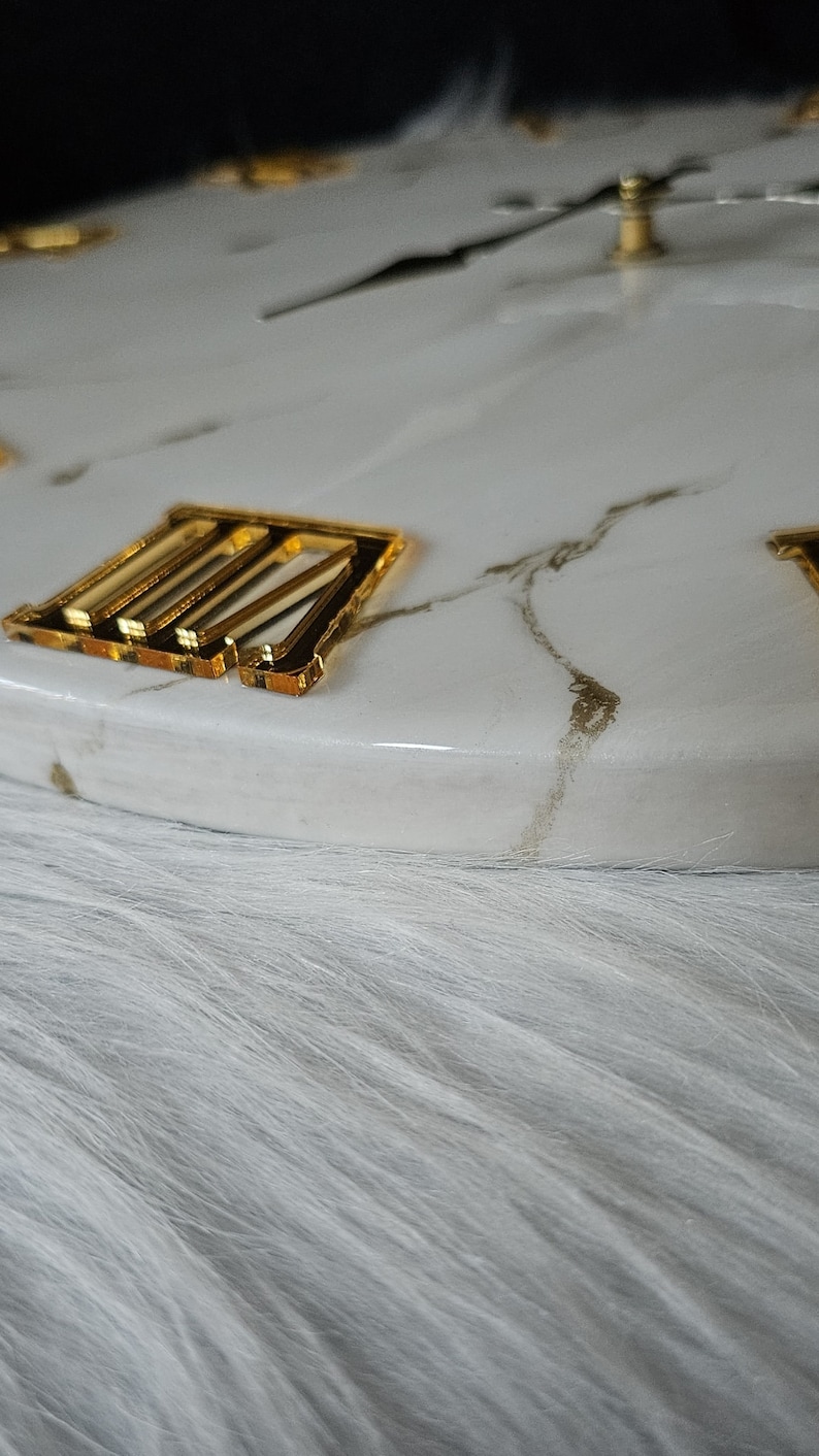May include: A white marble clock face with gold Roman numeral markings. The clock face is on a white fluffy surface.
