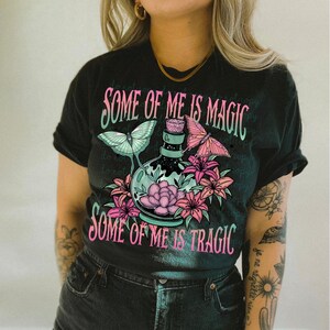 May include: Black t-shirt with a graphic of a potion bottle surrounded by flowers and butterflies. The text "Some of me is magic" and "Some of me is tragic" is printed on the shirt.