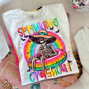 Puede incluir: Camiseta blanca con un gr&aacute;fico colorido de un esqueleto con gafas de sol y un sombrero para el sol, flotando en una boya inflable de arco&iacute;ris. La camiseta tiene las palabras "Sprinkling Crashout" en letras brillantes.