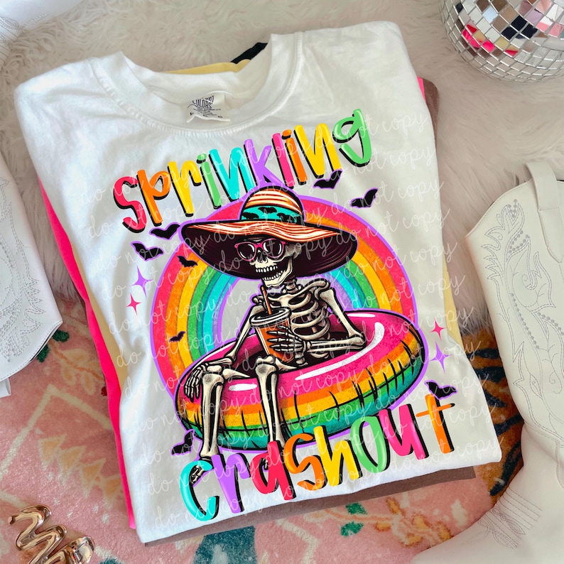 Puede incluir: Camiseta blanca con un gr&aacute;fico colorido de un esqueleto con gafas de sol y un sombrero para el sol, flotando en una boya inflable de arco&iacute;ris. La camiseta tiene las palabras "Sprinkling Crashout" en letras brillantes.