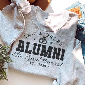 Puede incluir: Sudadera gris con texto negro que dice "LAW & ORDER ALUMNI Elite Squad Universit EST. 1999" con un gráfico de esposas.