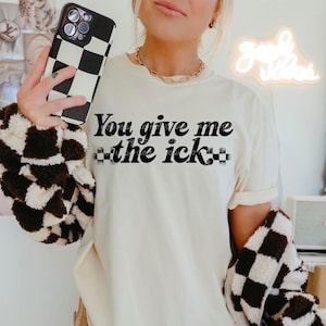 Puede incluir: Camiseta color crema con la frase "You give me the ick" en letras negras. La palabra "ick" está estilizada con un patrón de cuadros. La persona sostiene una funda de teléfono a cuadros en blanco y negro y lleva una chaqueta a juego.