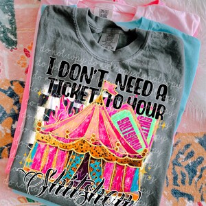以下が含まれることがあります： 「I DON'T NEED A TICKET TO YOUR SHITSHOW」の文字とカラフルなサーカステントのグラフィックがプリントされたグレーのComfort Colors Tシャツ。グレーのシャツの後ろには、ピンクとブルーのTシャツが重ねられています。
