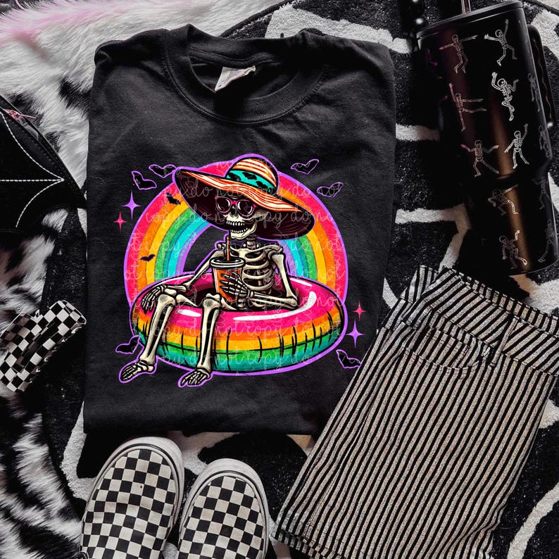 Puede incluir: Camiseta negra de cuello redondo con un dise&ntilde;o de esqueleto que lleva un sombrero para el sol y gafas de sol, relaj&aacute;ndose en un flotador inflable de arco&iacute;ris y sosteniendo una bebida. La camiseta se combina con pantalones cortos a rayas, zapatos a cuadros y un vaso con tem&aacute;tica de esqueleto.