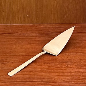 Op de afbeelding: Een zilveren taartschep met een driehoekig blad en een lange, rechthoekige handgreep. De schep is gemaakt van glanzend metaal en staat op een houten achtergrond. De schep is ontworpen voor het serveren van taarten.