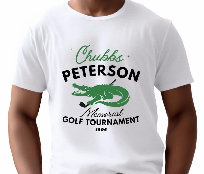 May include: White t-shirt with a green and black graphic. The graphic features a green alligator with a golf club and the words "Chubbs Peterson Memorial Golf Tournament 1996". The text is in black and green.