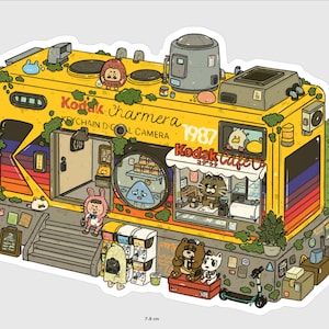 Op de afbeelding: Een gele sticker in de vorm van een camera met de tekst "Kodak Charmera" en "1987". De camera heeft een café met een kat-barista en diverse cartoonfiguren. De sticker meet 5,7 cm x 7,8 cm.