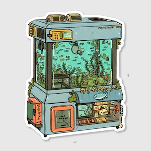 Könnte beinhalten: Ein blauer und orangefarbener Greifautomat mit einem großen Aquarium, gefüllt mit bunten Fischen, Pflanzen und Steinen. Die Maschine hat ein Schild mit der Aufschrift "カワイイ フィッシュ" in Japanisch. Die Maschine ist 5,7 cm hoch und 5 cm breit.