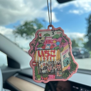 May include: A pink and white car air freshener shaped like a milk carton with a strawberry on top. The carton has a Japanese storefront with the text "いちご" and "カフェ" on the front. The storefront has a green awning and a window with two cartoon bunnies inside.