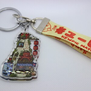 May include: A clear acrylic keychain featuring a colourful illustration of a Japanese street scene with a red octopus, a vending machine, and red lanterns. The keychain is attached to a yellow strap with the text "Kewpie Mayo" in red.