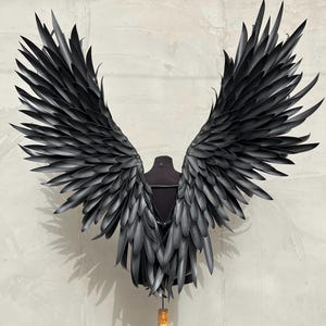 Puede incluir: Un par de alas de ángel grandes, negras y emplumadas. Las alas están hechas de plumas individuales que están superpuestas y superpuestas. Las alas están montadas en un soporte de madera.