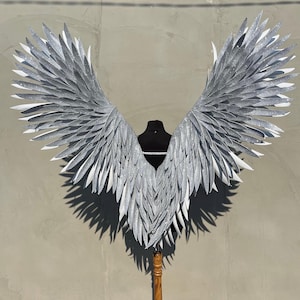 Puede incluir: Un par de alas de ángel grandes, de color plateado con purpurina, con una base blanca. Las alas están sujetas a un soporte de madera.