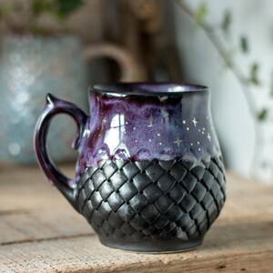 Pode incluir: Uma caneca de cerâmica com um design roxo escuro e preto. A metade superior da caneca tem um padrão de céu estrelado, enquanto a metade inferior apresenta uma textura tecida. A alça é de cor roxa escura.
