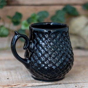 Può includere: Una tazza in ceramica nera con un design testurizzato a forma di squame. La tazza ha un'unica maniglia curva.