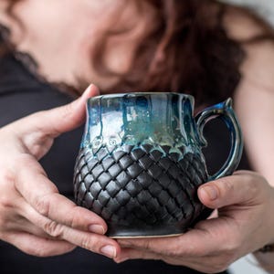 Peut inclure: Une tasse en céramique faite à la main avec un motif d'écailles de dragon unique. La tasse présente une palette de couleurs bleu foncé et noir avec un motif texturé sur la moitié inférieure. La poignée a un détail décoratif en forme d'aileron.