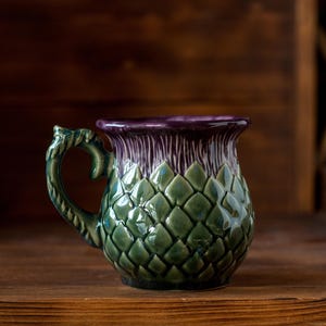 May include: A ceramic mug shaped like a thistle, featuring a deep purple top section with vertical ridges and a green base covered in overlapping, scale-like leaf patterns. The handle is sculpted to resemble a textured, twisted plant stem.