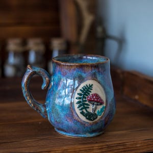 Könnte beinhalten: Ein Keramikbecher mit glänzender blau-violetter Glasur und einem erhabenen ovalen Medaillon auf der Vorderseite. Das Medaillon zeigt einen geformten grünen Farn und einen roten Pilz mit weißen Punkten. Der Becher hat einen geschwungenen Henkel.