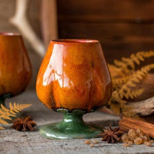 May include: Two ceramic goblets with a glossy, mottled orange finish resembling a pumpkin shape, each featuring a solid green pedestal base. The cups have a rounded, bulbous body and a smooth, reflective glaze that highlights the vibrant orange colour.