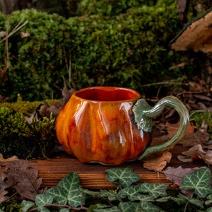 May include: A ceramic pumpkin-shaped mug with a green handle. The mug is glazed in shades of orange and brown, with a glossy finish. It is sitting on a wooden surface with green leaves.
