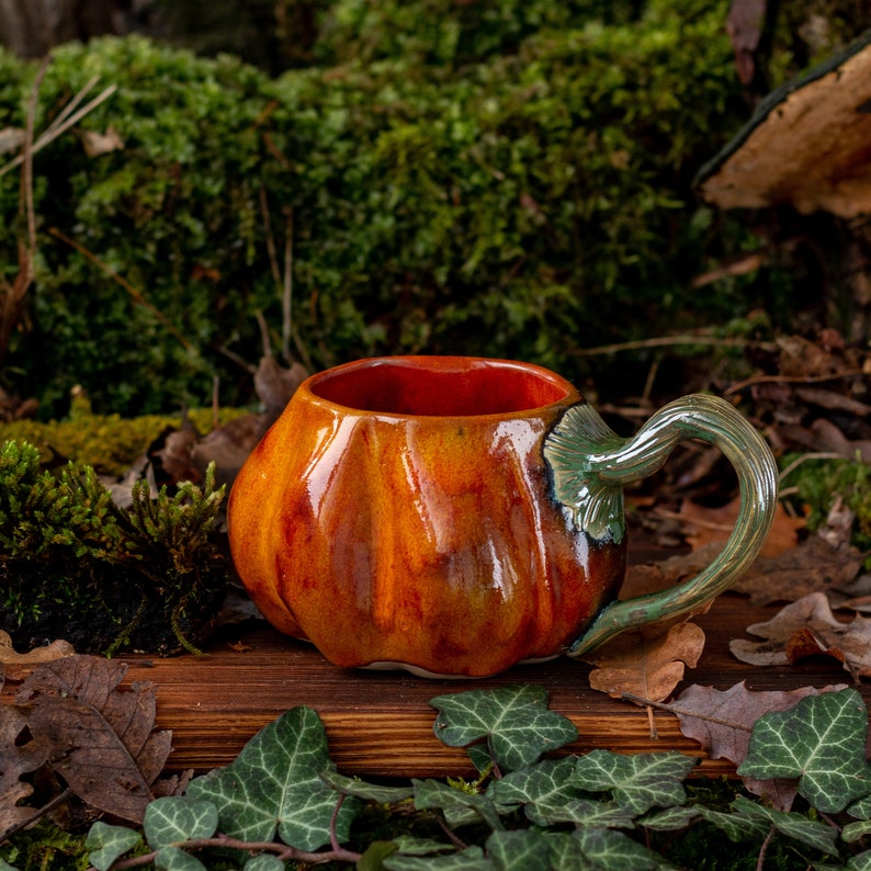 May include: A ceramic pumpkin-shaped mug with a green handle. The mug is glazed in shades of orange and brown, with a glossy finish. It is sitting on a wooden surface with green leaves.