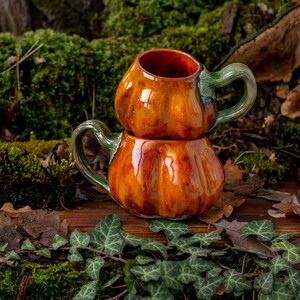 May include: Two stacked ceramic pumpkin mugs with green glaze accents. The mugs are sitting on a wooden surface with moss and leaves.