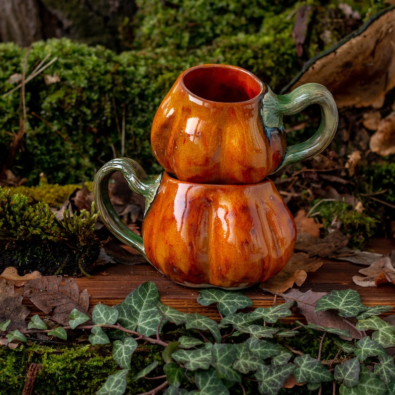 May include: Two stacked ceramic pumpkin mugs with green glaze accents. The mugs are sitting on a wooden surface with moss and leaves.
