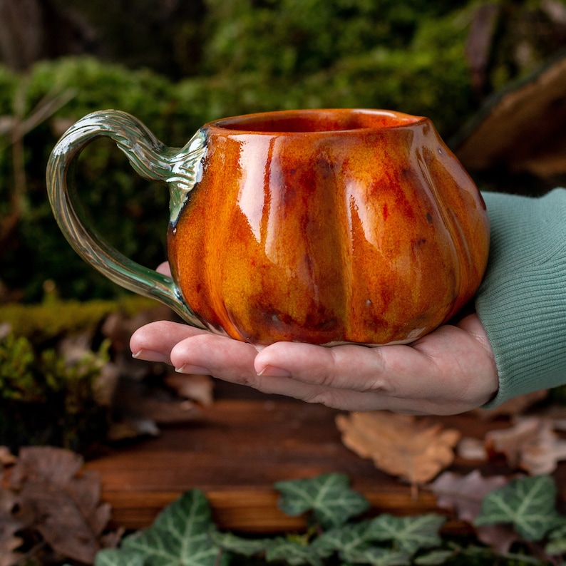 May include: A ceramic pumpkin-shaped mug with a green handle. The mug is glazed in shades of orange and brown.