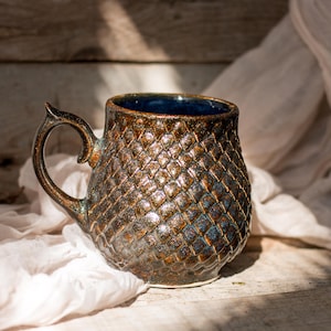 Może przedstawiać: Brązowy kubek ceramiczny z niebieską glazurą i fakturowanym wzorem w kształcie rombu. Kubek ma unikalny, zakrzywiony uchwyt.