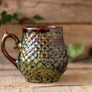 May include: A green and brown ceramic mug with a textured, scale-like design. The mug has a rounded body and a handle.