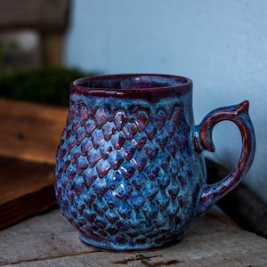 Puede incluir: Taza de cerámica hecha a mano con un patrón texturizado en forma de escamas en tonos azules y burdeos. La taza tiene un asa única y un acabado brillante. Ideal para café o té.