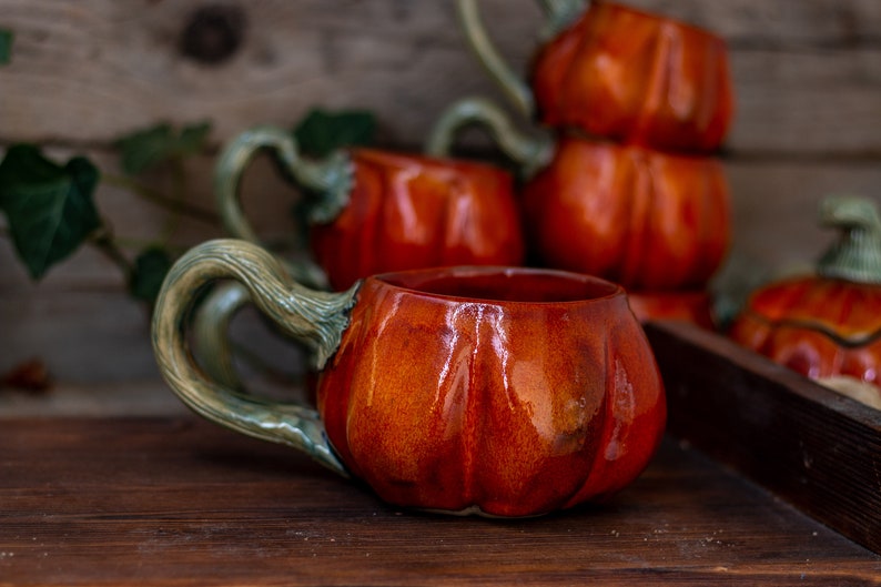 May include: A set of four ceramic pumpkin mugs with green handles. The mugs are a deep orange color and have a glossy finish.