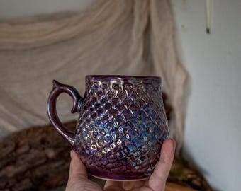 May include: A handcrafted, iridescent ceramic mug with a textured, scale-like pattern. The mug features a deep purple hue with hints of blue and green, and a curved handle. This unique mug is perfect for coffee or tea.