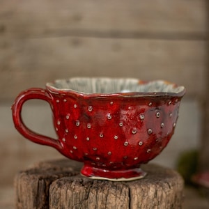 Może przedstawiać: Czerwony ceramiczny kubek do herbaty z białymi plamkami i falistym brzegiem. Kubek ma uchwyt i stoi na drewnianej powierzchni.