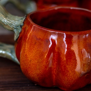 May include: A ceramic pumpkin-shaped mug with a green handle. The mug is glazed in a deep orange color with a glossy finish.